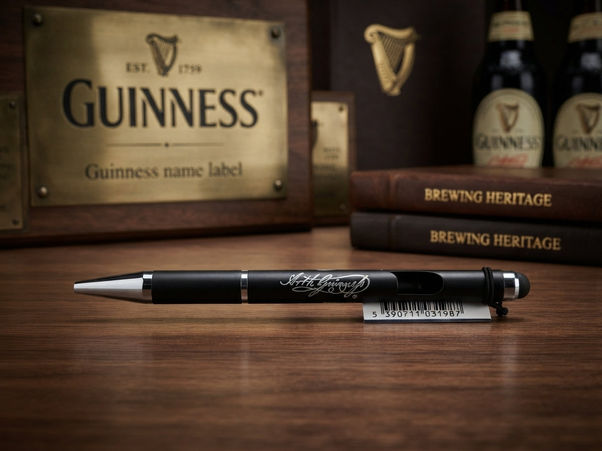 A black and silver Guinness-branded pen resting on a wooden tabletop, with a branded Guinness plaque, books, and bottles blurred in the background.
